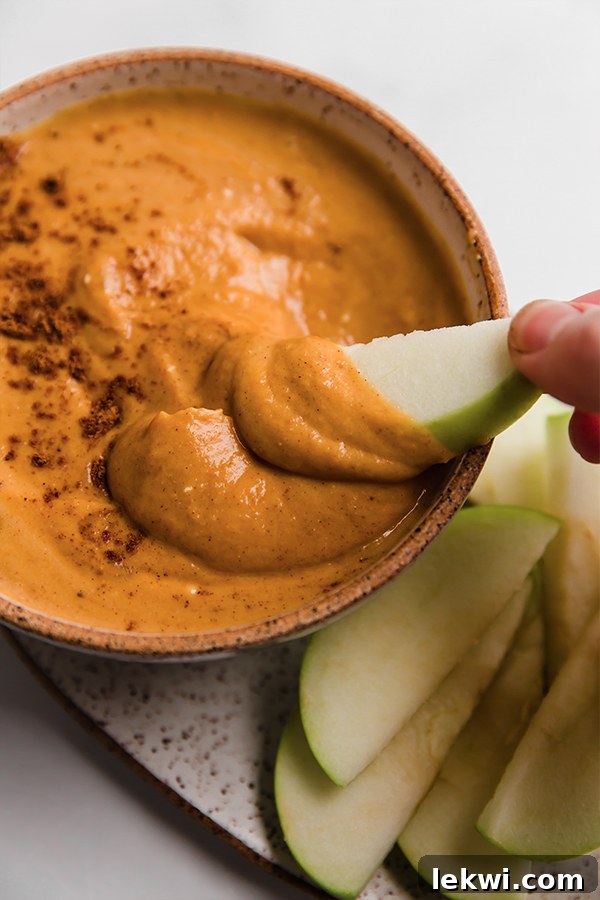 A vibrant green apple slice, perfectly coated in the rich, creamy pumpkin pie dip, being lifted from a rustic bowl. The image highlights the dip's luxurious texture and its ideal pairing with fresh fruit, inviting viewers to take a bite.