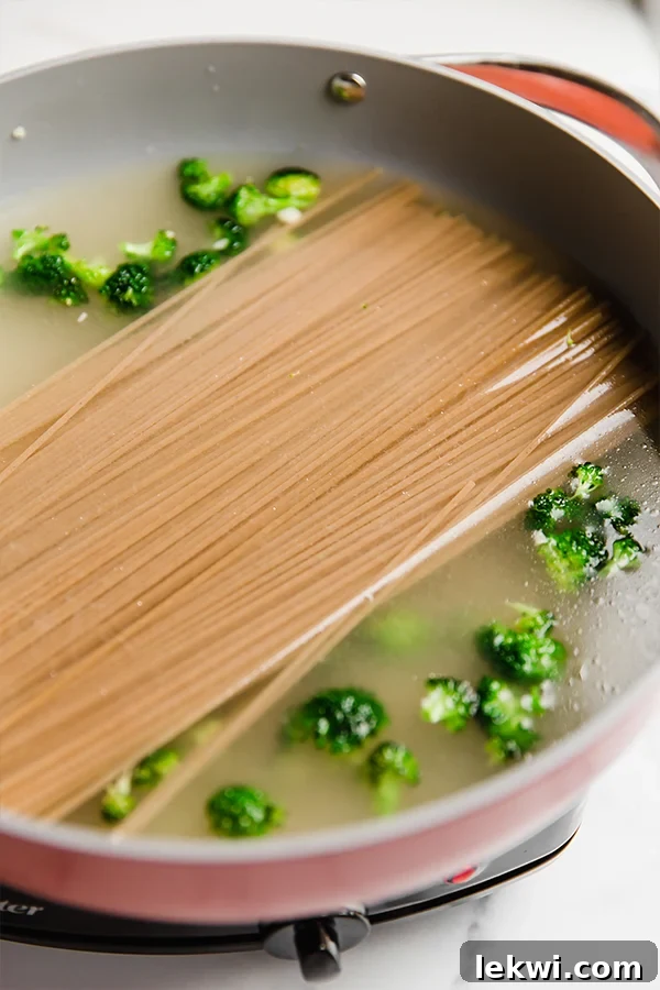 A large, deep pan filled with uncooked spaghetti, broccoli, and broth, ready for cooking.
