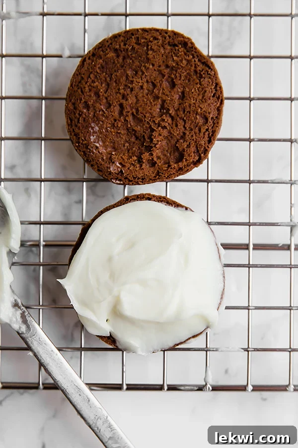 An open-face chocolate cookie being generously iced with white peppermint cream filling, ready to be sandwiched.