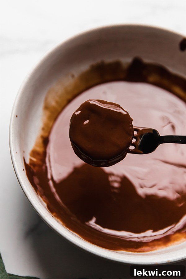 A chocolate sandwich cookie being dipped into a bowl of melted chocolate with a fork, showing the smooth coating process.