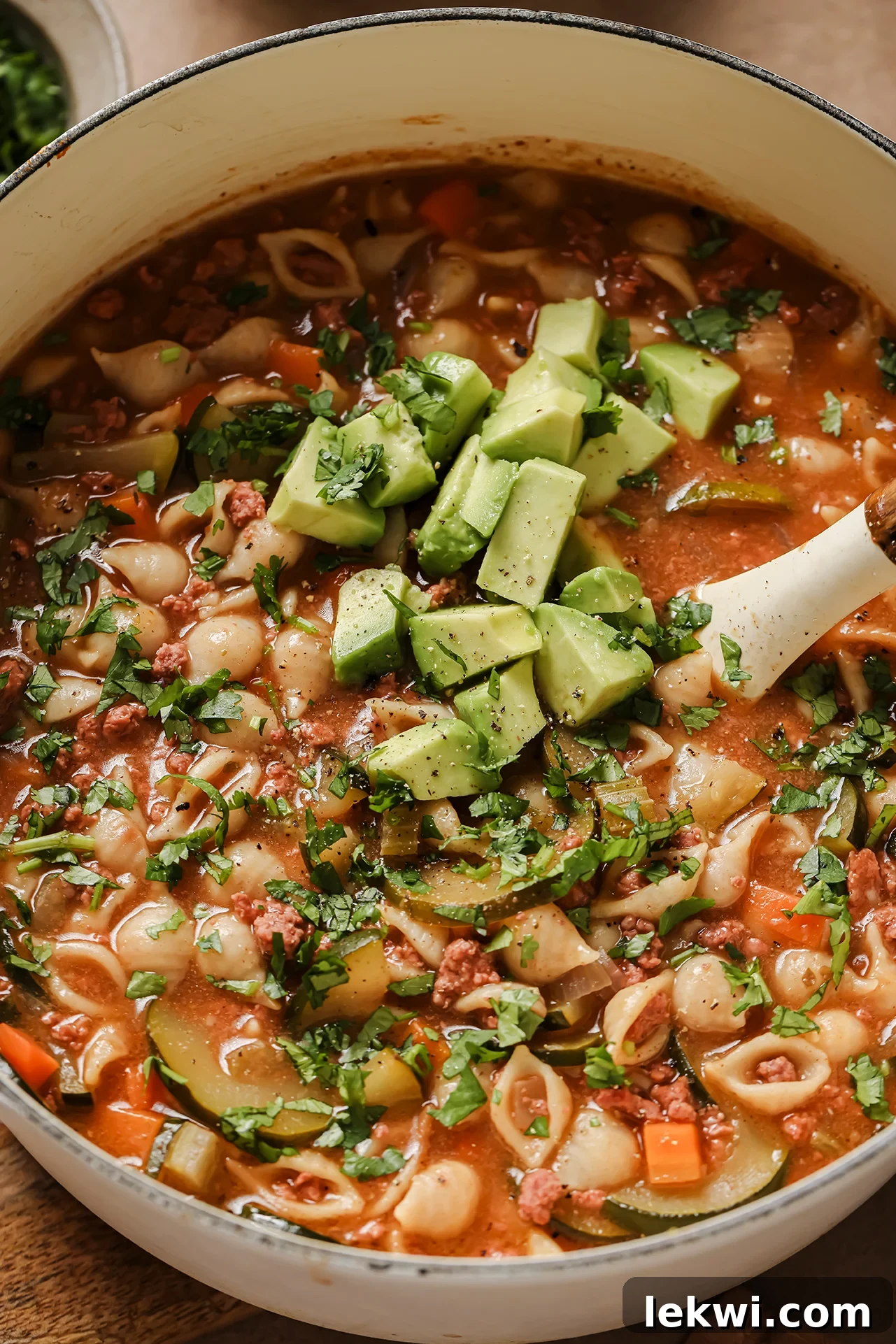 A large white Dutch oven filled with rich Conchitas con Carne soup, generously topped with fresh avocado slices and cilantro, ready to be served.