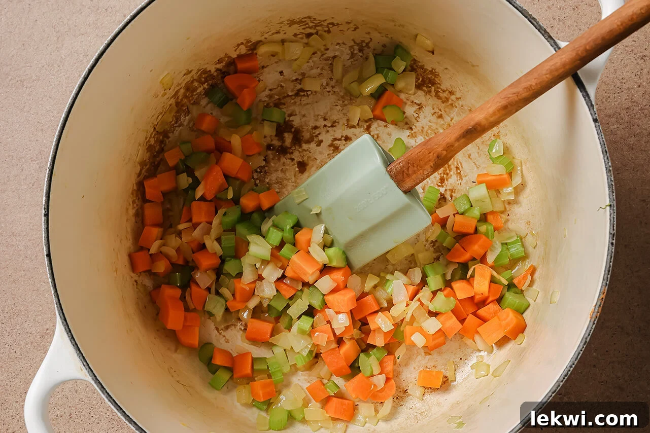 Diced onion, celery, and carrots gently cooking in a white Dutch oven, forming the aromatic base of the soup.