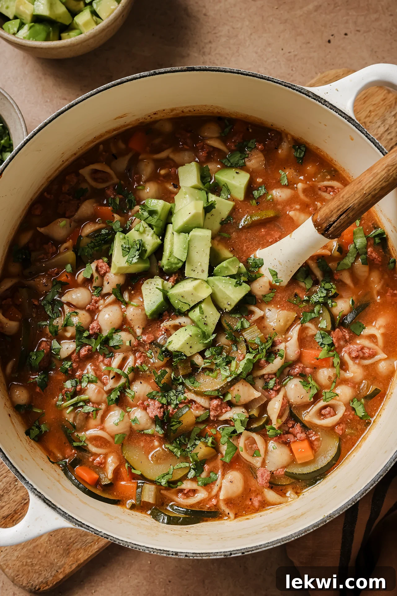 A large white Dutch oven brimming with hot Conchitas con Carne soup, beautifully garnished with fresh avocado slices and cilantro, with extra avocado on the side.