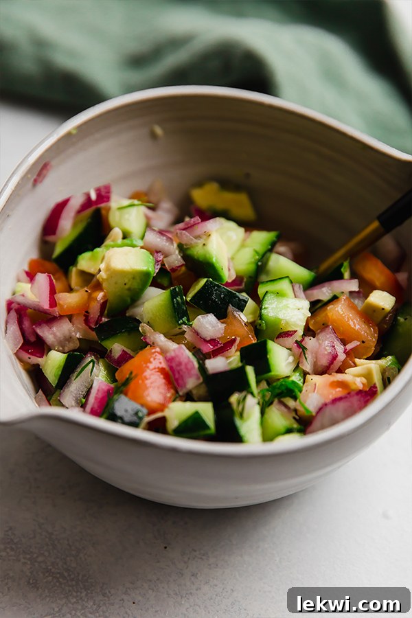 A vibrant, colorful bowl of freshly chopped vegetables for Greek salsa, featuring cucumbers, tomatoes, red onions, and avocado.