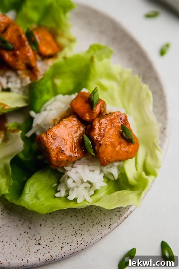 A beautifully presented plate featuring several firecracker salmon lettuce cups. The golden-brown salmon pieces, coated in a rich red sauce, are served in crisp butter lettuce leaves, garnished with fresh green onions and a sprinkle of sesame seeds. The image conveys a sense of freshness and deliciousness.