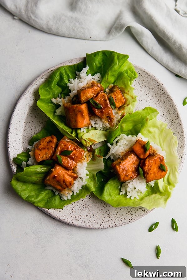 A plate featuring three perfectly assembled firecracker salmon lettuce cups. Each cup is filled with saucy salmon and rice, topped with green onions, and resting on a white ceramic plate against a light background.