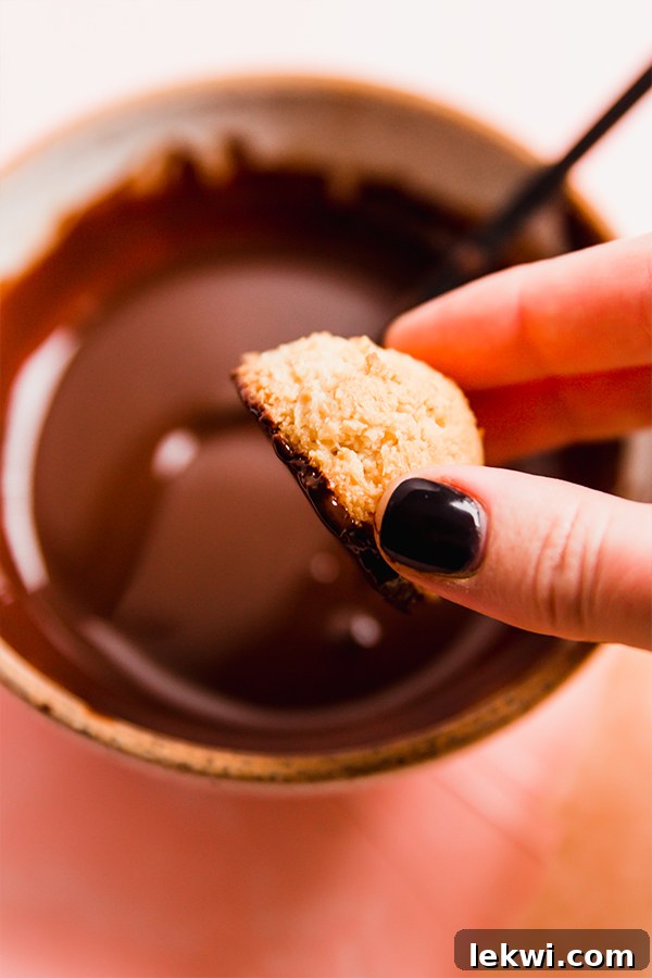 A hand dipping a baked coconut macaroon into a bowl of melted dairy-free chocolate, showing the process of coating.
