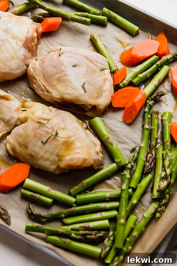 A sheet pan loaded with marinated chicken thighs, bright green asparagus spears, chopped carrots, and vibrant orange slices, ready to be roasted in the oven.