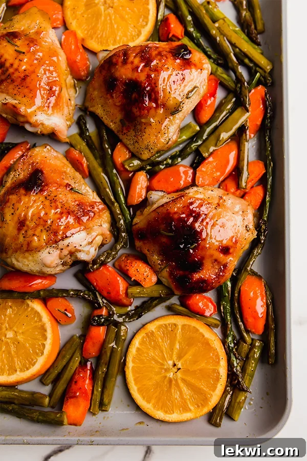 Sheet pan filled with perfectly roasted citrus glazed chicken thighs and a colorful array of vegetables like asparagus, carrots, and bright orange slices, all glistening with a sweet and tangy glaze.