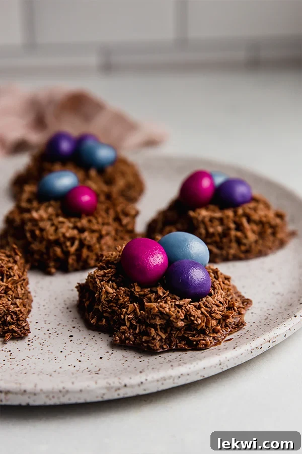 A cluster of beautifully formed chocolate coconut Easter egg nests, each adorned with colorful candy gems, presented on a white plate.