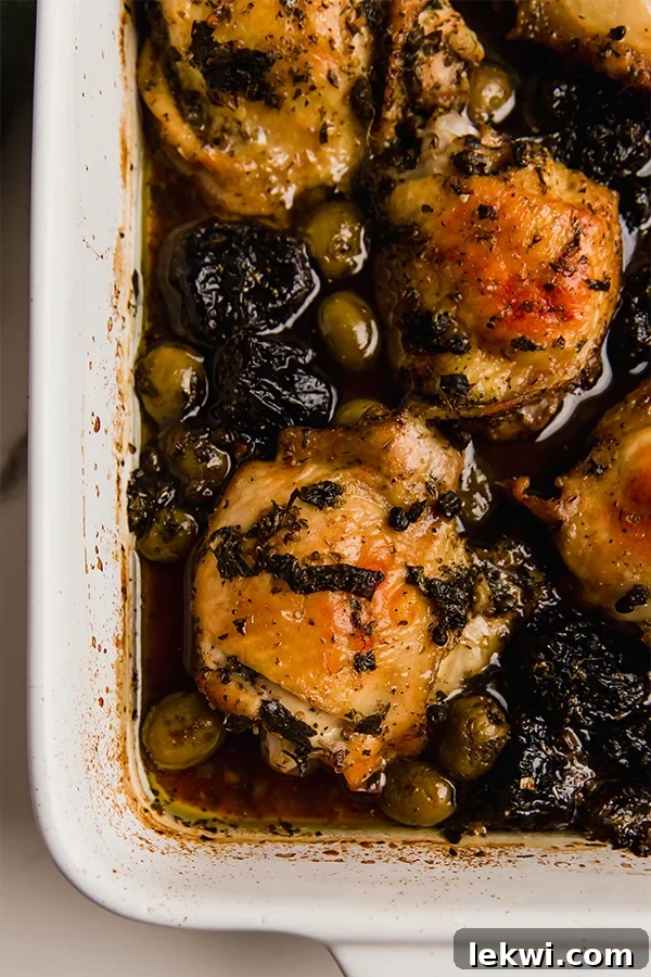 Chicken Marbella baking in a casserole dish, bubbling with its rich sauce of olives, capers, and prunes, topped with glistening chicken thighs.