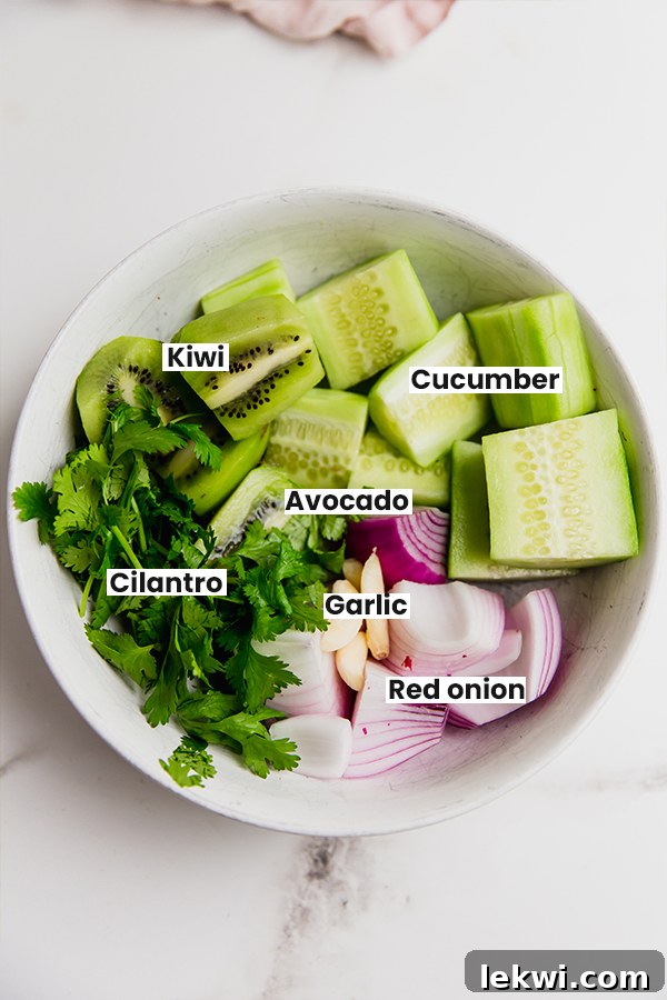 An array of fresh ingredients for AIP Nightshade-Free Salsa Verde including cucumber, kiwi, avocado, onions, and herbs, laid out on a cutting board.