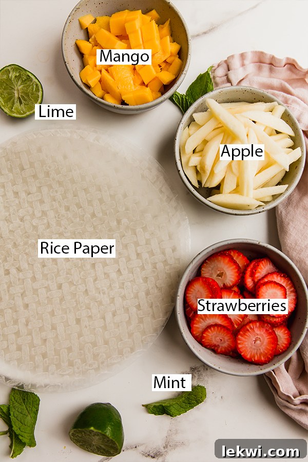 A colorful array of ingredients for fruit spring rolls, including rice paper, mint, sliced strawberries, diced mango, and julienned apple.