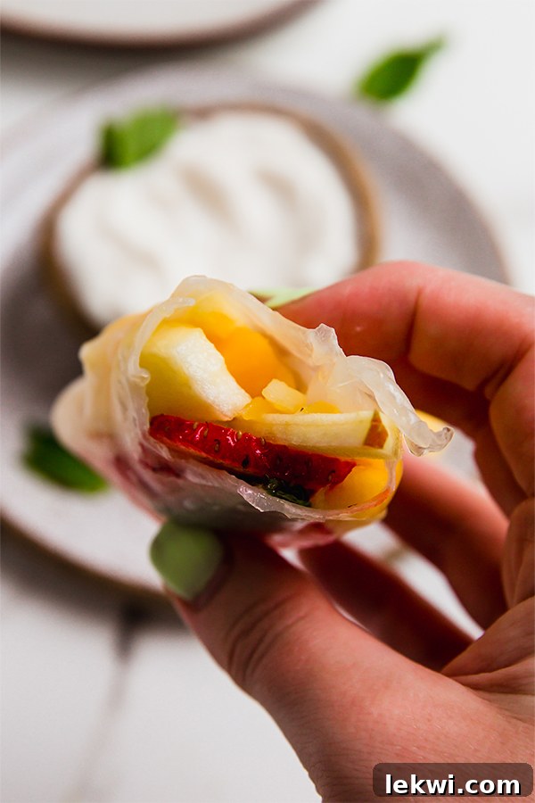 A fruit spring roll with a bite taken out, revealing the colorful layers of fruit and mint inside, ready to be dipped.
