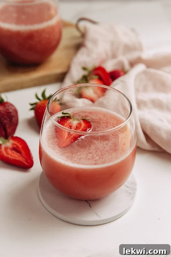 A chilled glass of vibrant pink homemade Frosé, garnished with a fresh strawberry slice on the rim, sits invitingly on a bright background, hinting at summer refreshment.