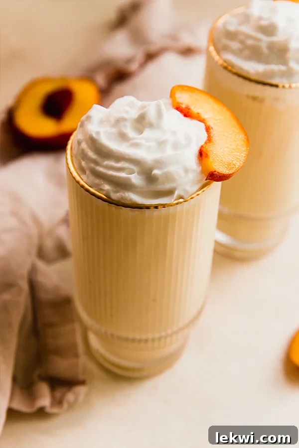 Tall glass filled with a creamy dairy-free peach milkshake, garnished with a fresh peach slice on the rim.