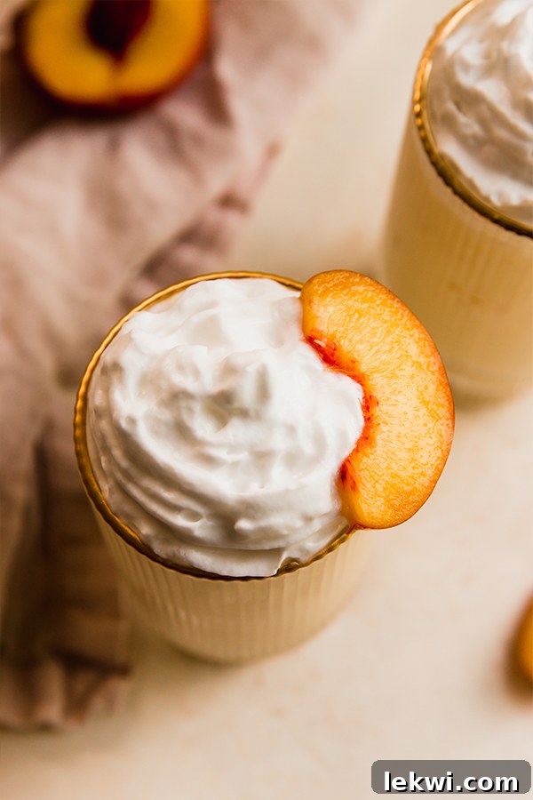 Close-up of a dairy-free creamy peach milkshake topped with a generous swirl of coconut whipped cream and a fresh peach slice.