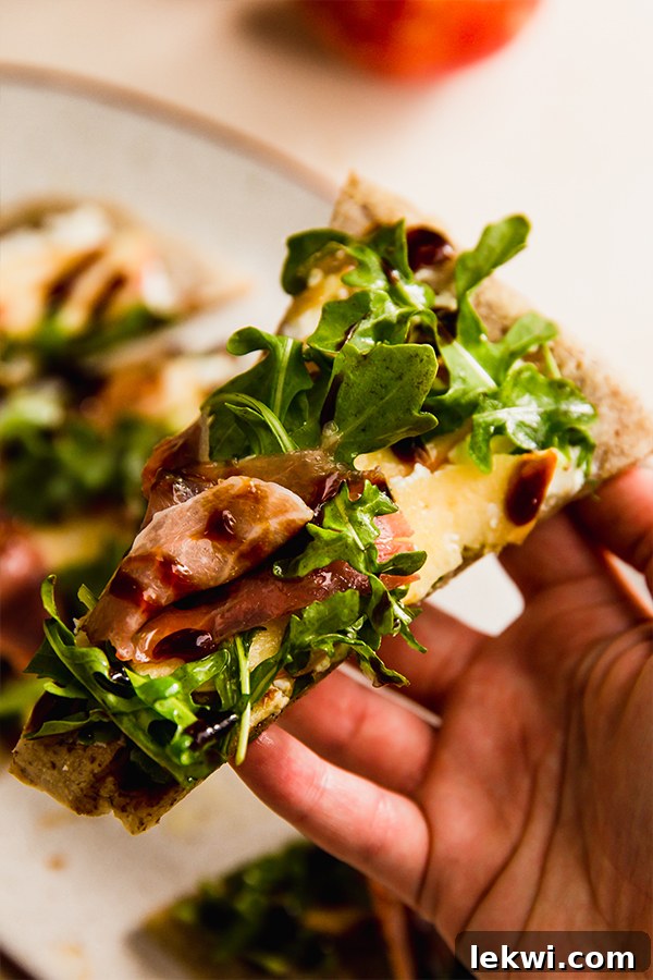 A slice of apple balsamic flatbread held in a hand, showcasing the crispy crust and fresh toppings.