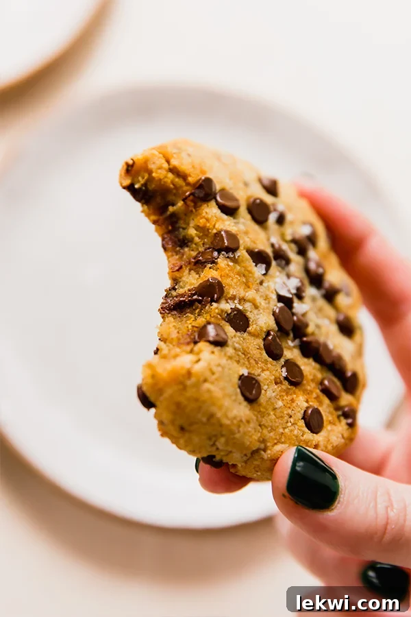 A person holding a large, golden-brown pumpkin chocolate chip cookie, with a bite taken out of it, showcasing its soft interior and melted chocolate.