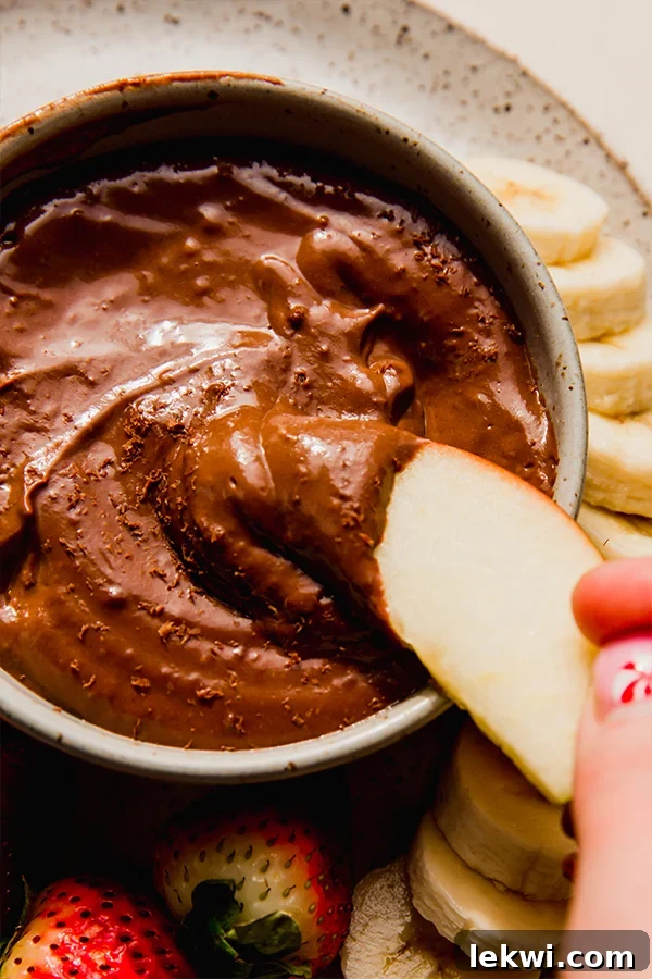 A sliced red apple being dipped into a bowl of creamy, dairy-free chocolate fruit dip.