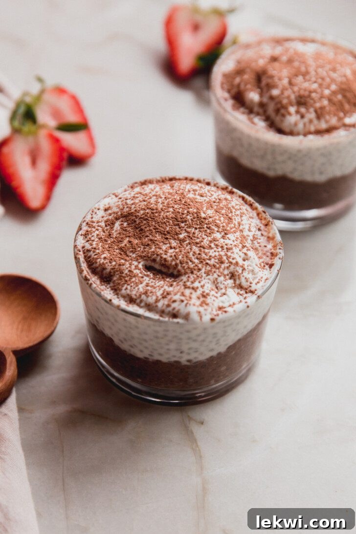 A small glass of Tiramisu Chia Pudding, with vibrant berries and fresh mint leaves artfully arranged in the background, creating an inviting and refreshing dessert scene.