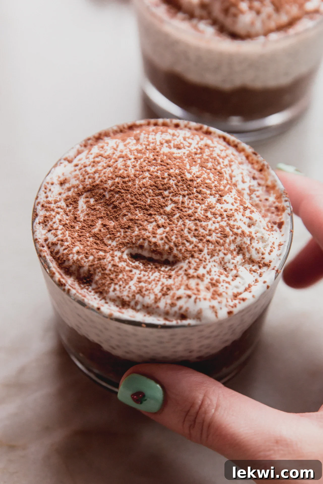 A hand holds up a clear glass cup filled with layered tiramisu chia pudding, showcasing its decadent chocolate and creamy 'mascarpone' layers, ready to be enjoyed.
