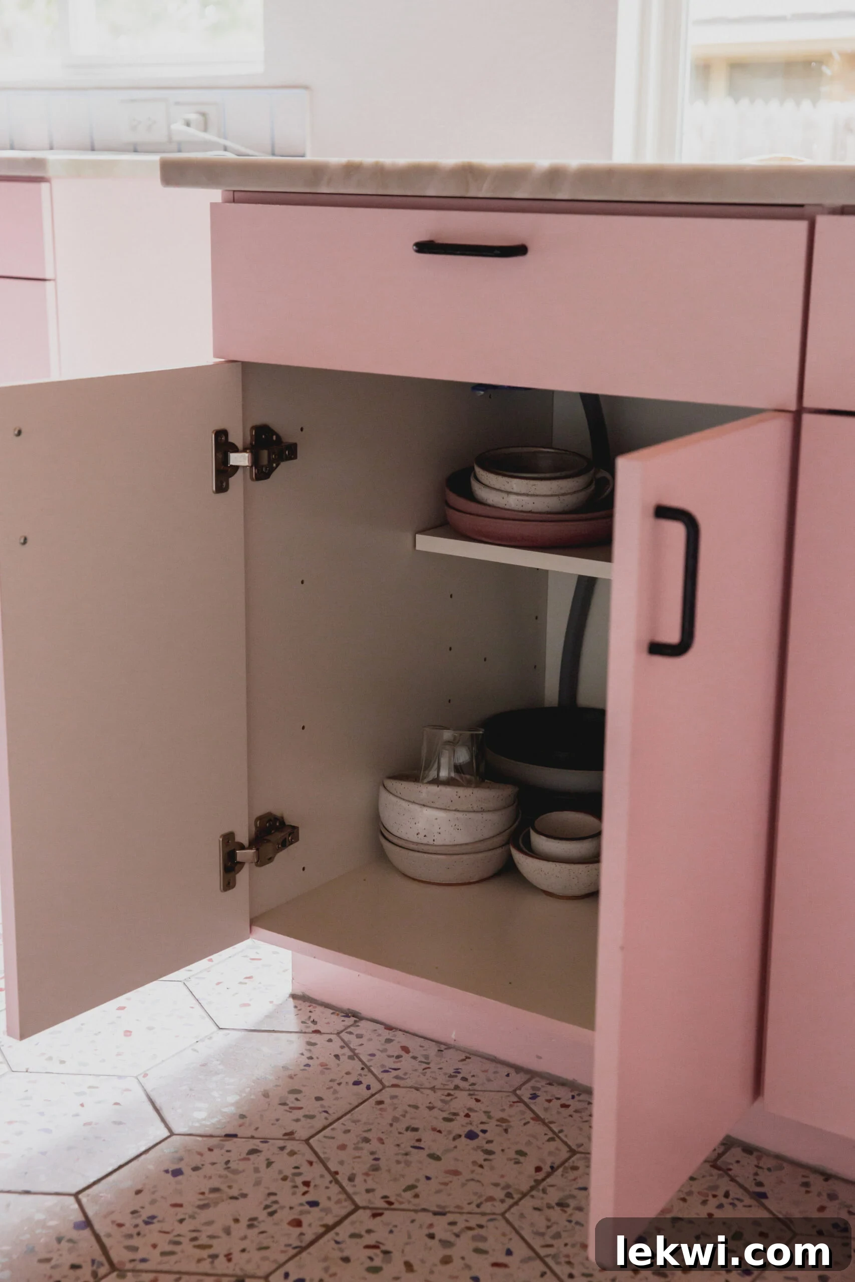 Open pink cabinet in the kitchen island, revealing organized dishes and photography props for a food blogger.