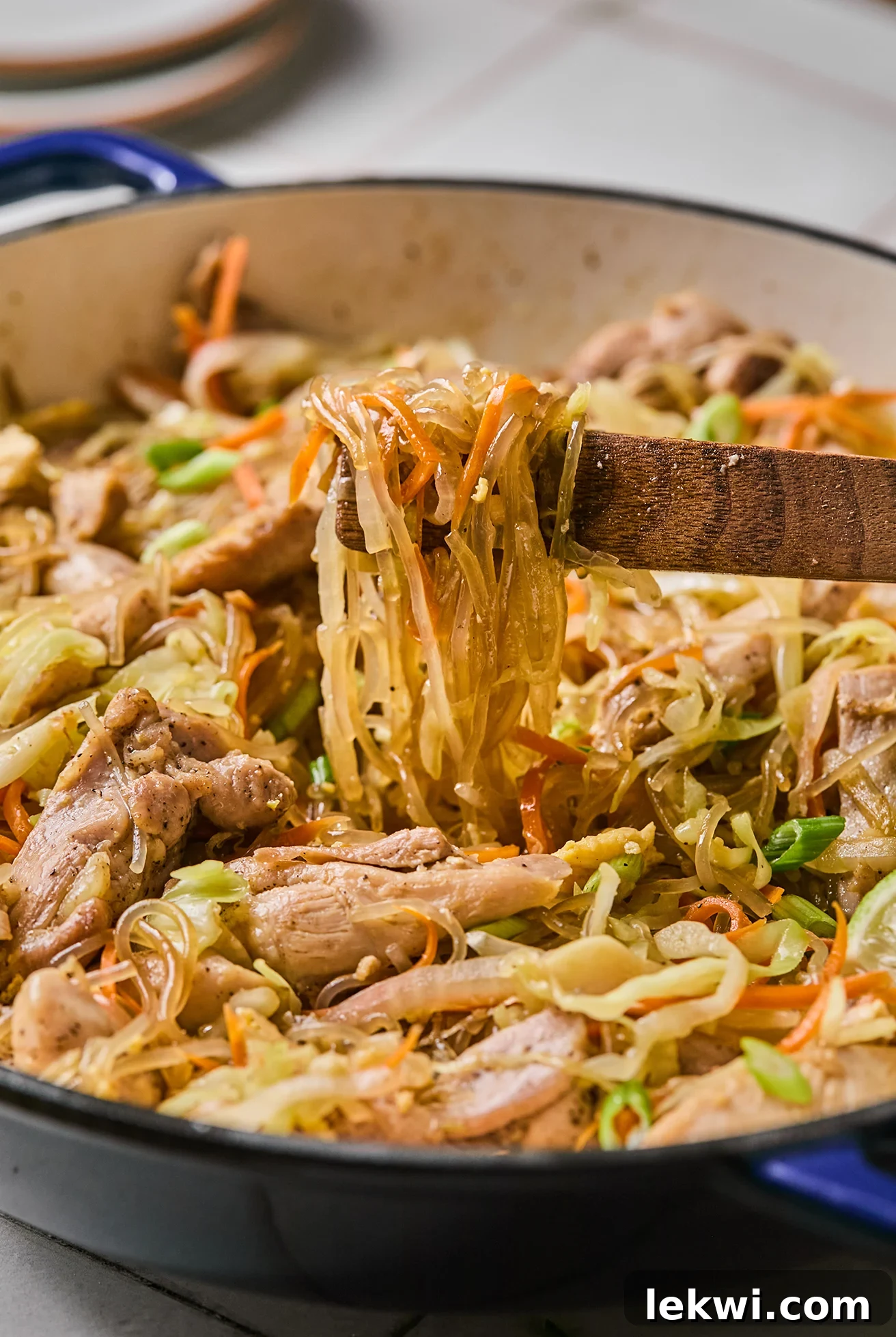 A large pan filled with Pad Woon Sen, showing the mix of glass noodles, chicken, and vegetables, with a pair of serving tongs ready.