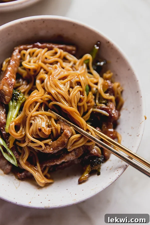 A close-up shot of a bowl filled with Sheet Pan Beef & Broccoli Ramen, featuring vibrant green onions as a garnish. A pair of chopsticks is poised to pick up a delicious bite.