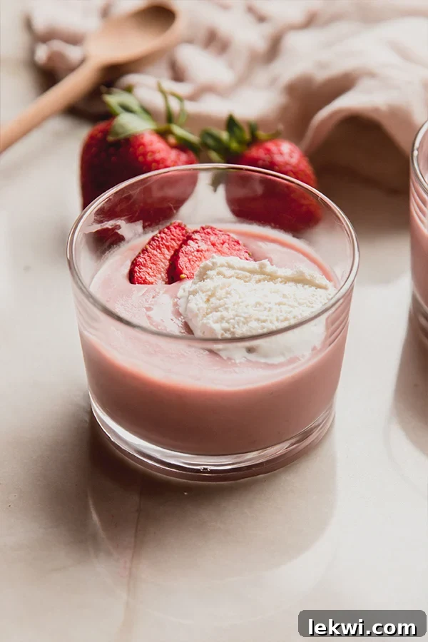 A beautifully presented cup of creamy strawberries and cream pudding, garnished with fresh strawberry slices, sits on a kitchen counter, awaiting a moment of pure enjoyment.