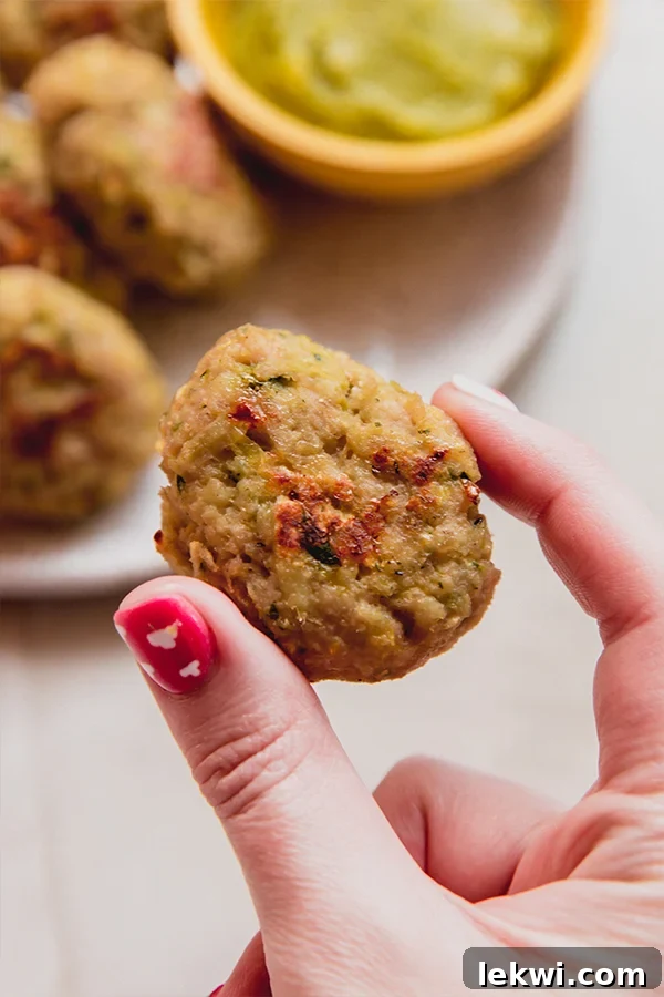 A close-up shot of a hand holding a single, golden-brown zucchini chicken nugget, showcasing its inviting texture and appetizing appearance. The detail highlights the successful crispness achieved during baking, making it irresistible.