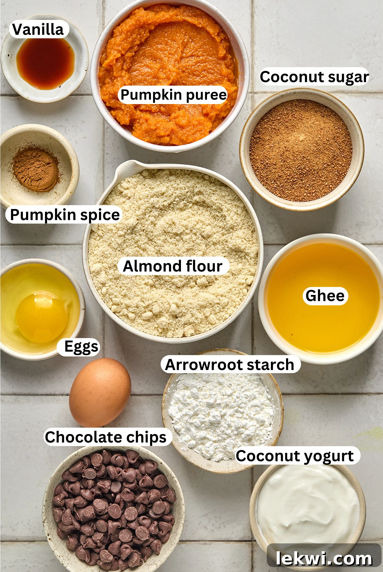 A curated display of ingredients for gluten-free pumpkin pie cookie bars, including pumpkin puree, almond flour, dairy-free chocolate chips, and coconut yogurt, all laid out in individual bowls.