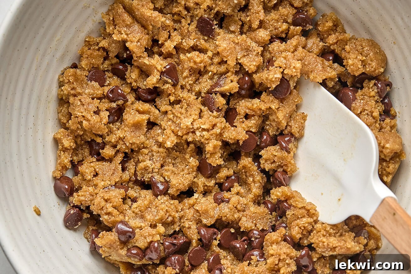 Wet ingredients for the cookie layer being mixed into the dry ingredients, forming a dough. Dairy-free chocolate chips are seen ready to be folded in.