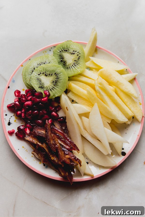 A beautifully arranged plate featuring thinly sliced kiwi, julienned apples, pears, dates, and scattered pomegranate arils, ready for assembling the fresh fruit rice paper rolls.