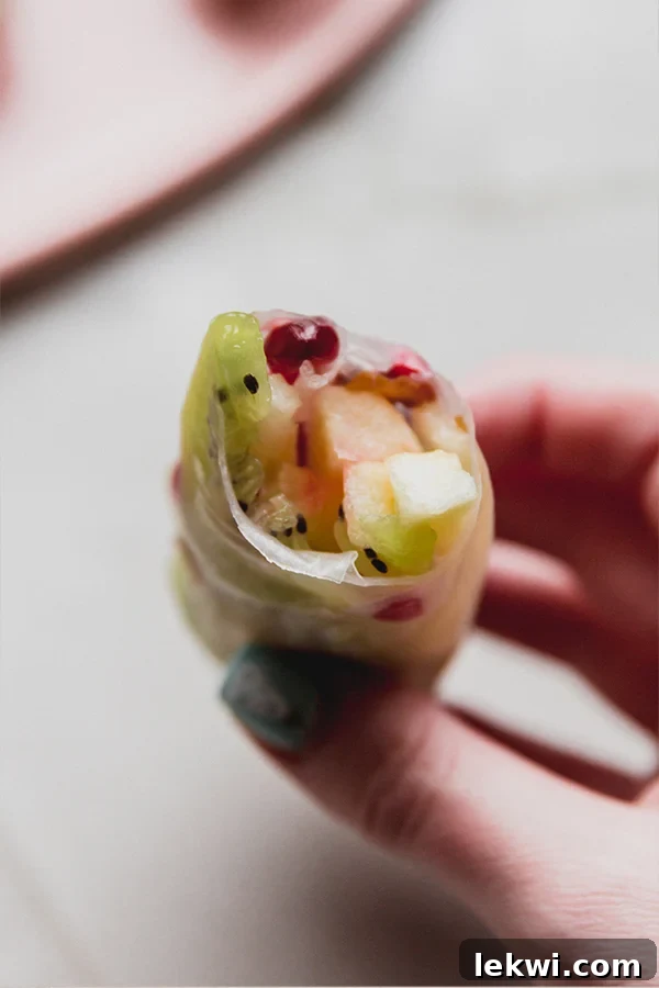 A vibrant winter fruit rice paper roll with a bite taken out, revealing the colorful kiwi, apple, pear, and pomegranate arils inside.