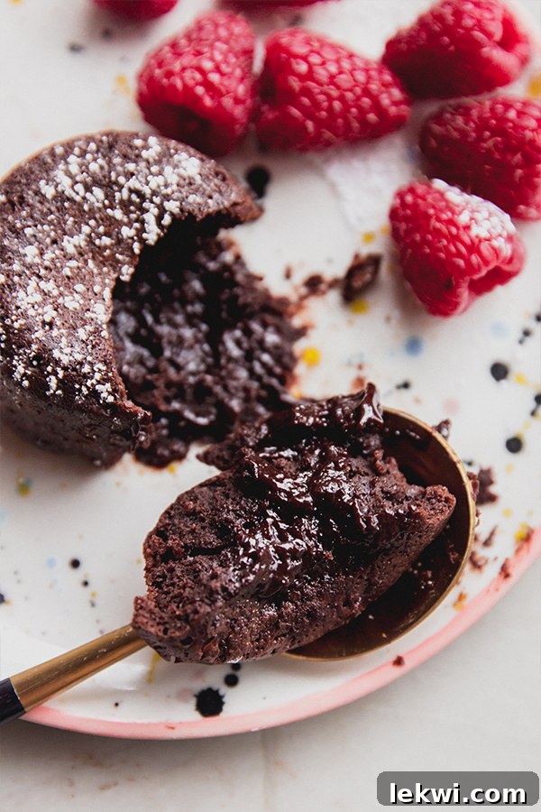 A gluten-free chocolate lava cake on a plate with a spoon digging into the molten center, surrounded by fresh raspberries.