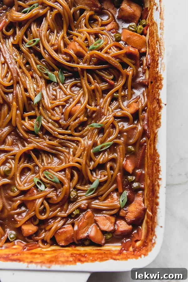 A vibrant baking dish filled with golden-brown baked honey garlic chicken noodles, ready to be served. The noodles are coated in a rich sauce, intermingled with tender chicken pieces and colorful vegetables.