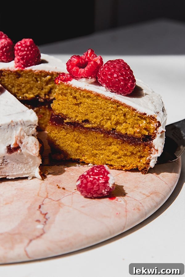 A perfectly sliced two-layer gluten-free white chocolate raspberry cake, revealing its vibrant raspberry filling, topped with fresh raspberries and a dusting of icing sugar.