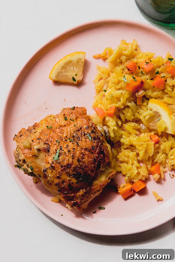 Overhead image of one Greek lemon chicken thigh on a pink plate with a rice and diced carrots on the side, topped with lemon wedges and a green glass with water on the side.