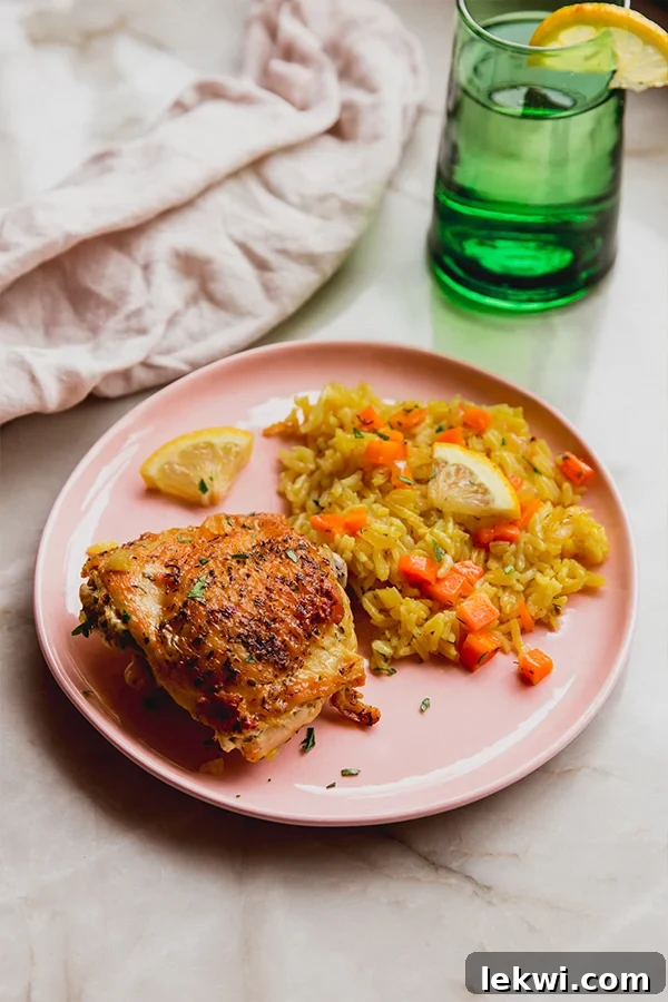 Greek lemon chicken on a pink plate with a rice and diced carrots on the side, topped with lemon wedges and a green glass with water on the side.