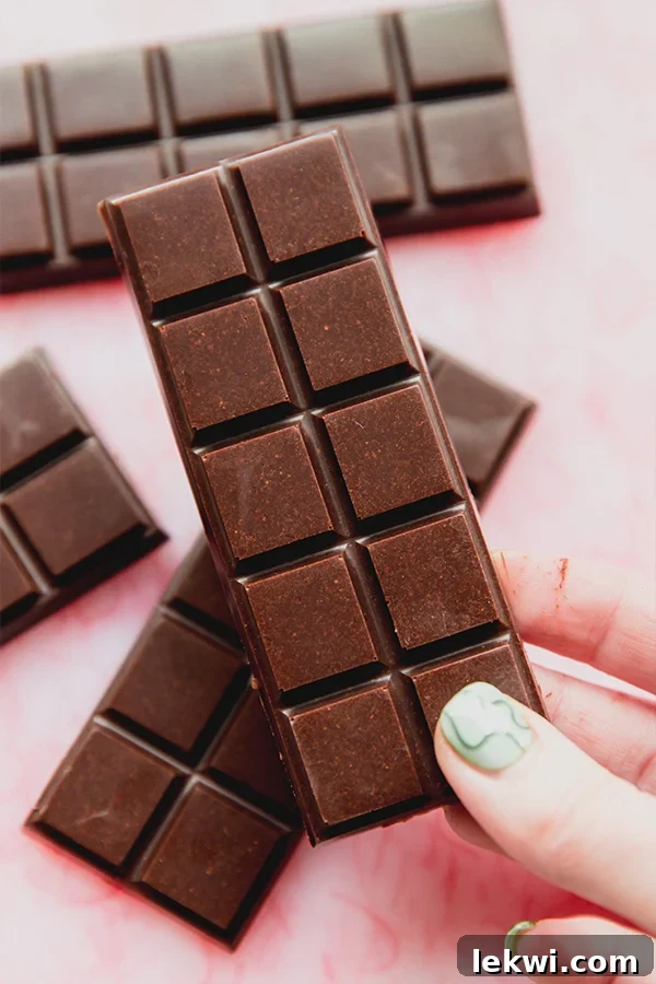 A hand holds a square of homemade coconut milk chocolate bar, with several full bars resting on a pink cutting board in the background, highlighting its delicious texture.