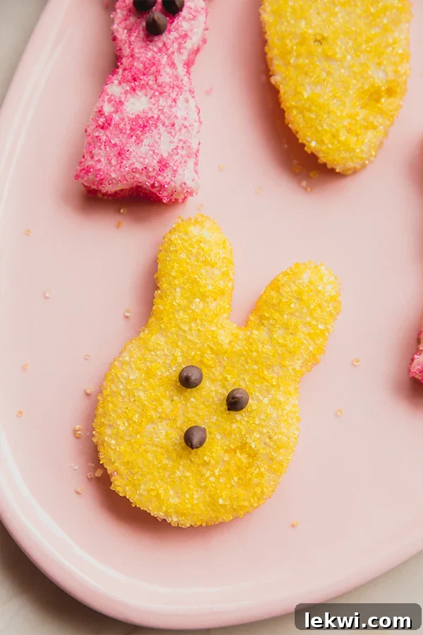 Bunny shaped marshmallow coated in yellow decorating sugar with three chocolate chips to make a face, sitting on a pink plate.