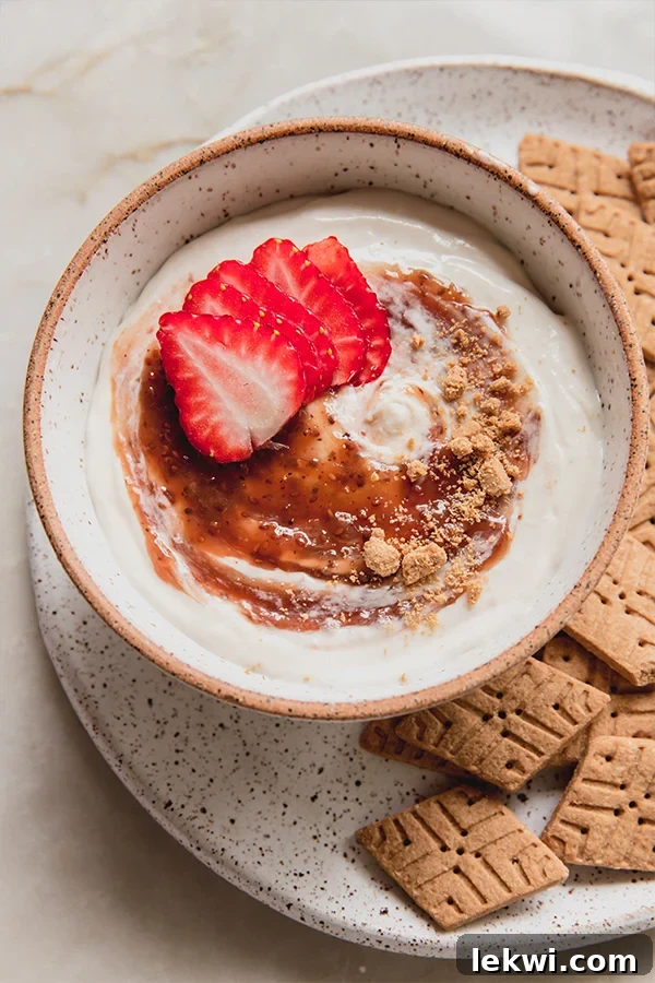 A vibrant bowl of dairy-free strawberry cheesecake dip, garnished with fresh strawberries and crushed gluten-free graham crackers, with whole crackers arranged invitingly on the side for dipping.