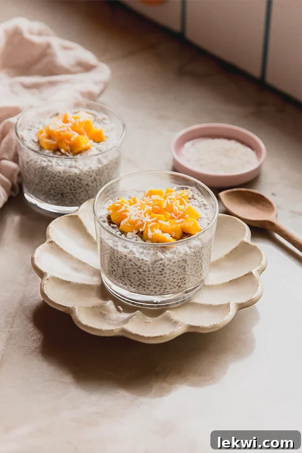 Two bowls of Mango Sticky Rice Chia Pudding. The front bowl is beautifully topped with fresh diced mango and shredded coconut, accompanied by a small wooden spoon, ready to be enjoyed. Another blurred bowl sits in the background, hinting at more deliciousness.