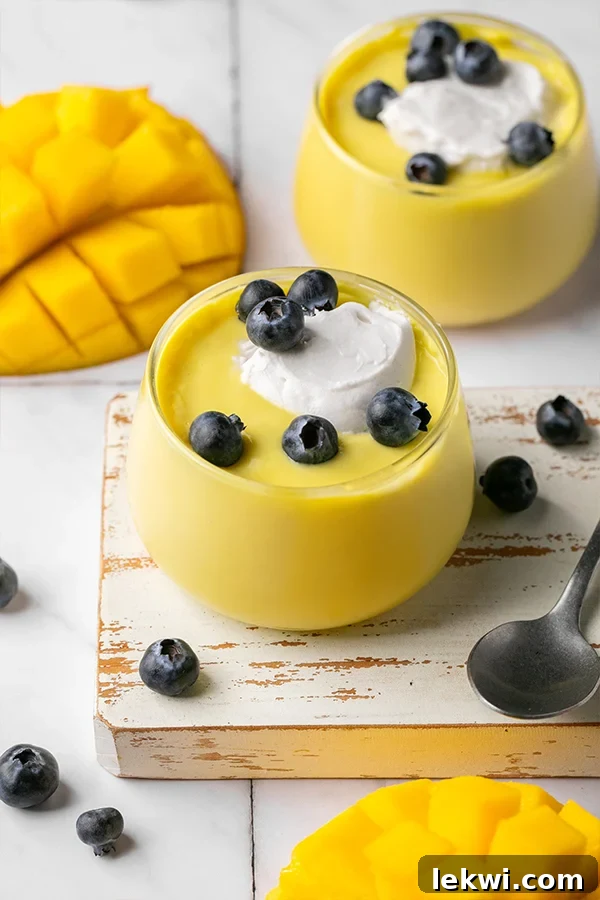 Mango pudding in a glass dish topped with coconut cream and blueberries, with a spoon on the side.