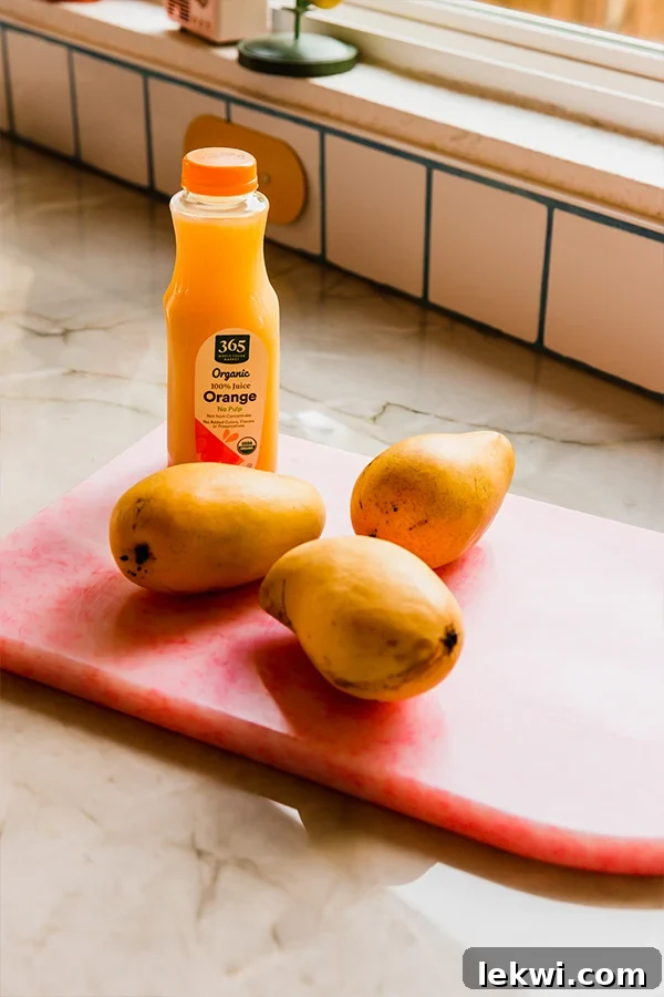 Three ripe Ataulfo mangoes and a carton of orange juice on a pink cutting board, showcasing the simple ingredients for sorbet.