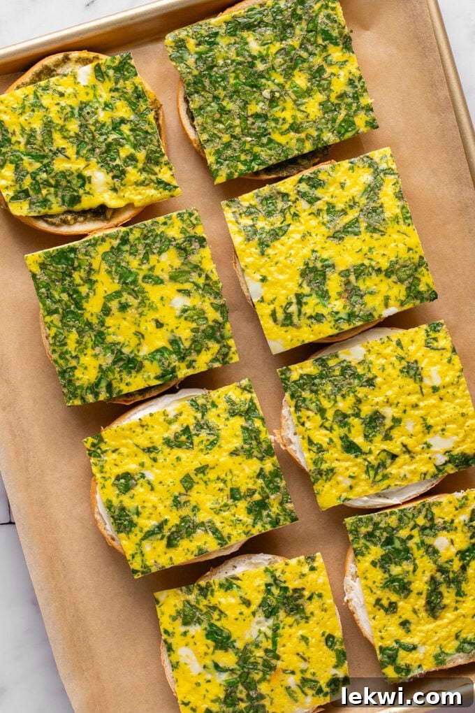 A parchment-lined baking sheet with prepared sheet pan egg slices, some topped with spinach, ready for breakfast sandwiches, placed in meal prep bags.
