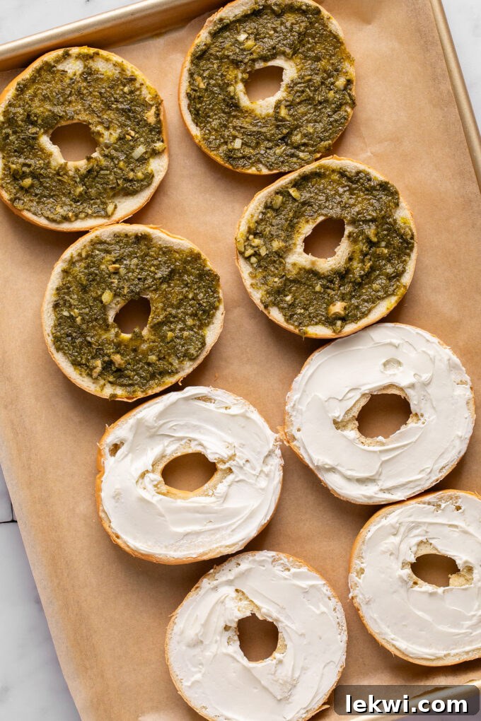 Halved bagels on a baking sheet, with some topped with cream cheese and others with pesto, ready for layering.