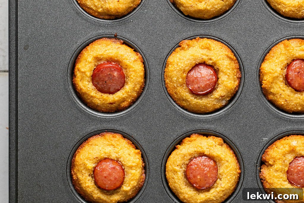 Freshly baked corn dog muffins, golden brown and puffed up, still in the muffin tin.