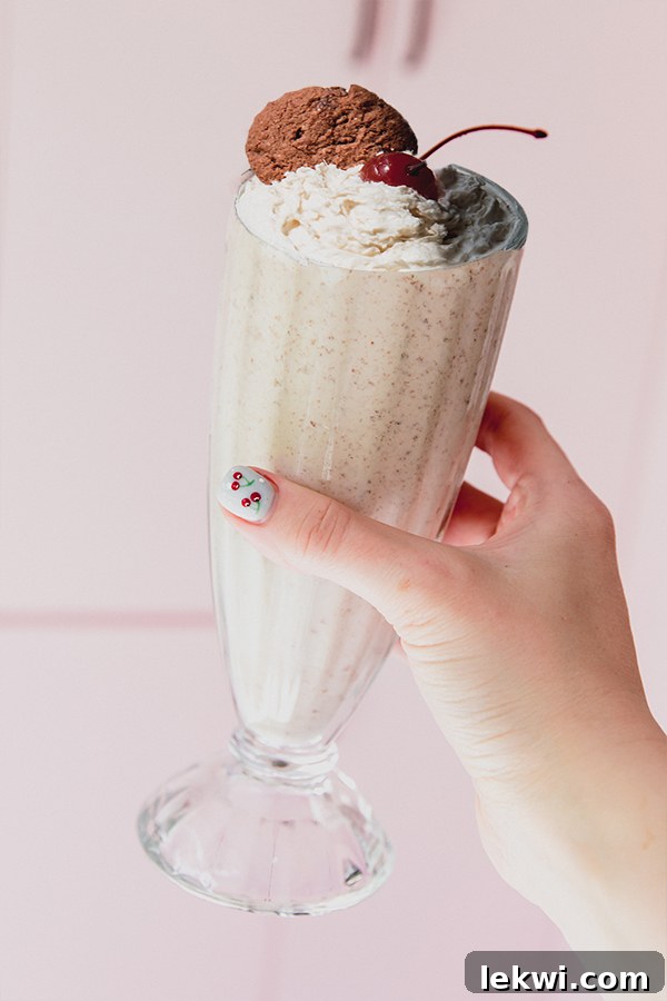 A hand holding a tall milkshake cup filled with a Ninja Creami cookies and cream milkshake, topped with whipped cream, a cookie, and a cherry.